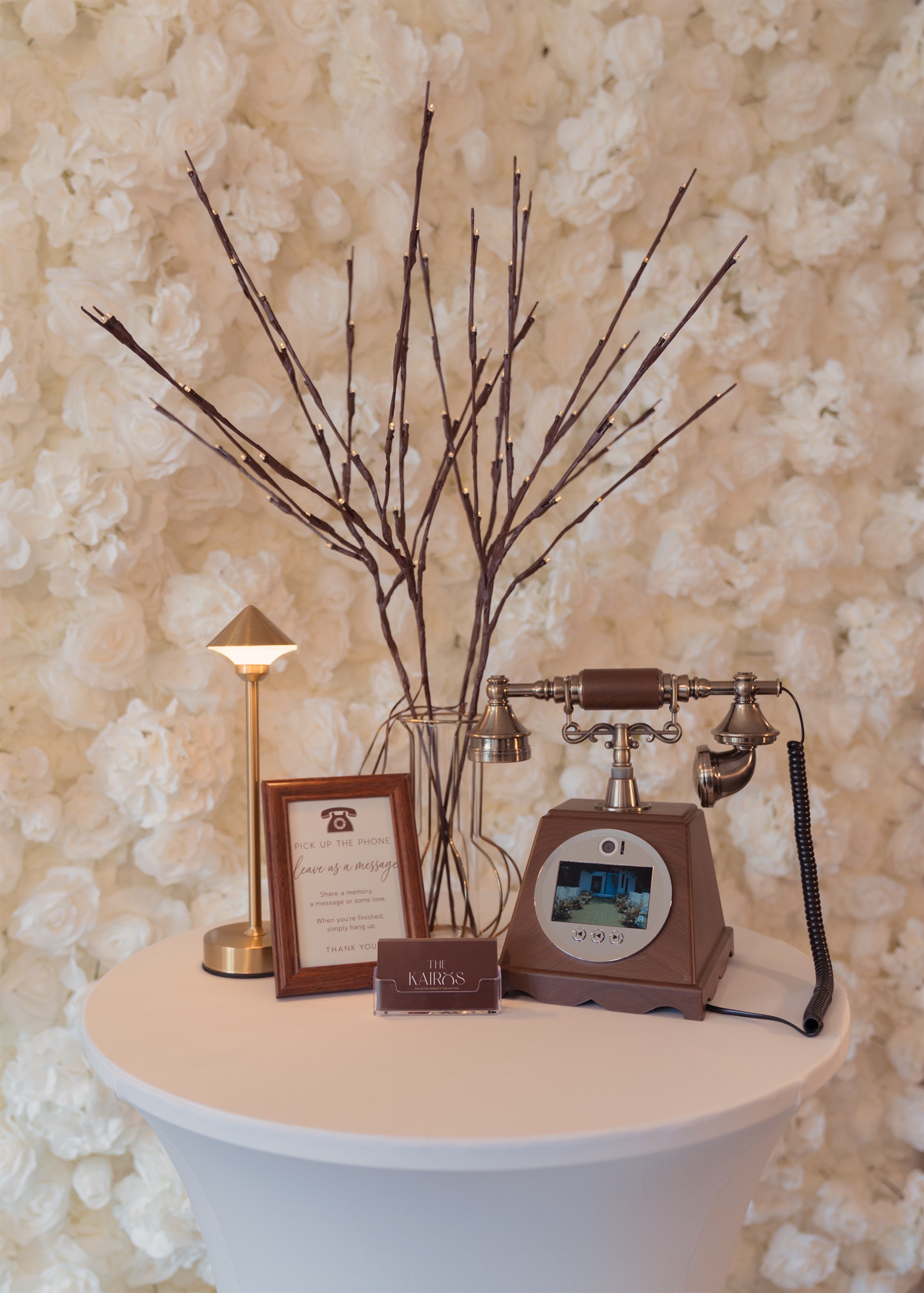 The Heirloom video and audio guestbook telephone on a white table with a gold lamp, framed sign and illuminated branches
